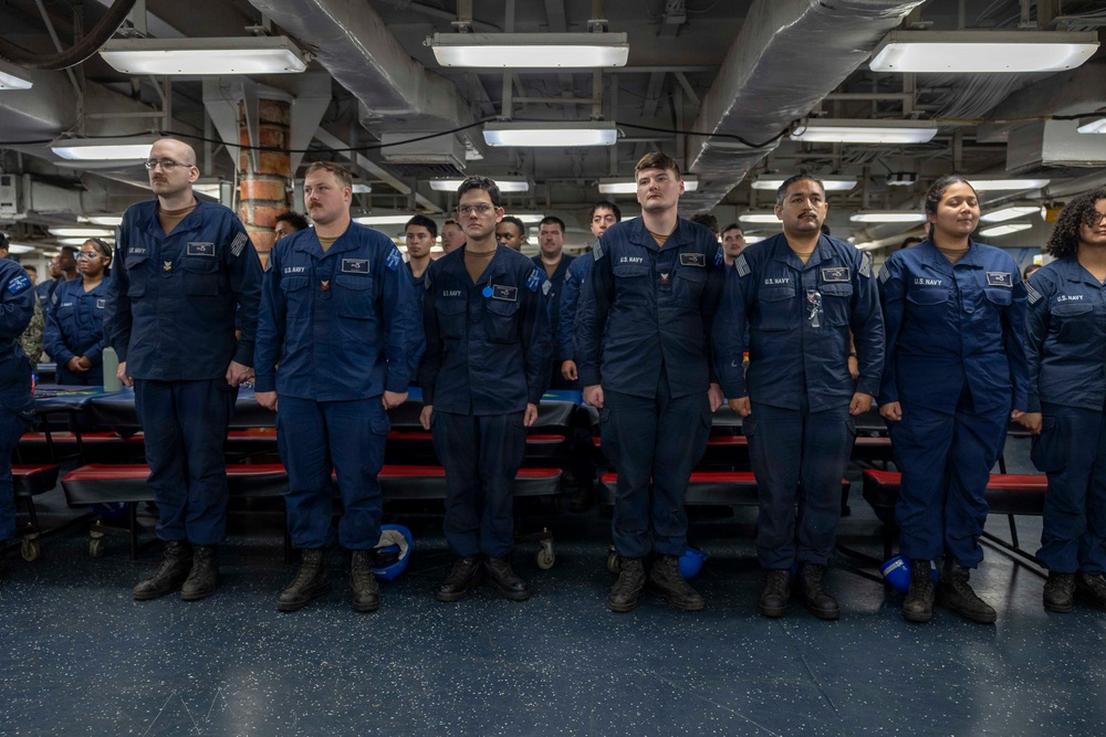 A group of U.S. Navy personnel in standard blue Navy Working Uniforms (NWUs) are standing at attention in what appears to be a mess deck or berthing area aboard a naval vessel. Multiple sailors are arranged in rows, facing forward in a formal formation. The setting is an interior shipboard space with overhead fluorescent lighting, pipes, and red-seated tables visible.