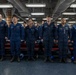 A group of U.S. Navy personnel in standard blue Navy Working Uniforms (NWUs) are standing at attention in what appears to be a mess deck or berthing area aboard a naval vessel. Multiple sailors are arranged in rows, facing forward in a formal formation. The setting is an interior shipboard space with overhead fluorescent lighting, pipes, and red-seated tables visible.