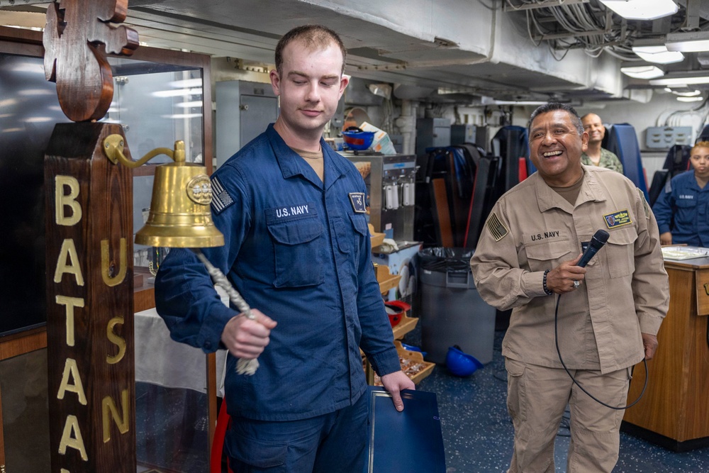 Two U.S. Navy personnel are standing near a ship's bell mounted on a wooden post bearing the name 'BATAN' (likely USS Bataan). The sailor in blue NWU Type III uniform is holding the bell's rope lanyard, while a senior sailor in tan/khaki uniform holds a microphone and smiles broadly. The scene appears to be an informal ceremony or event taking place in an interior compartment of a naval vessel.