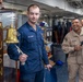 Two U.S. Navy personnel are standing near a ship's bell mounted on a wooden post bearing the name 'BATAN' (likely USS Bataan). The sailor in blue NWU Type III uniform is holding the bell's rope lanyard, while a senior sailor in tan/khaki uniform holds a microphone and smiles broadly. The scene appears to be an informal ceremony or event taking place in an interior compartment of a naval vessel.
