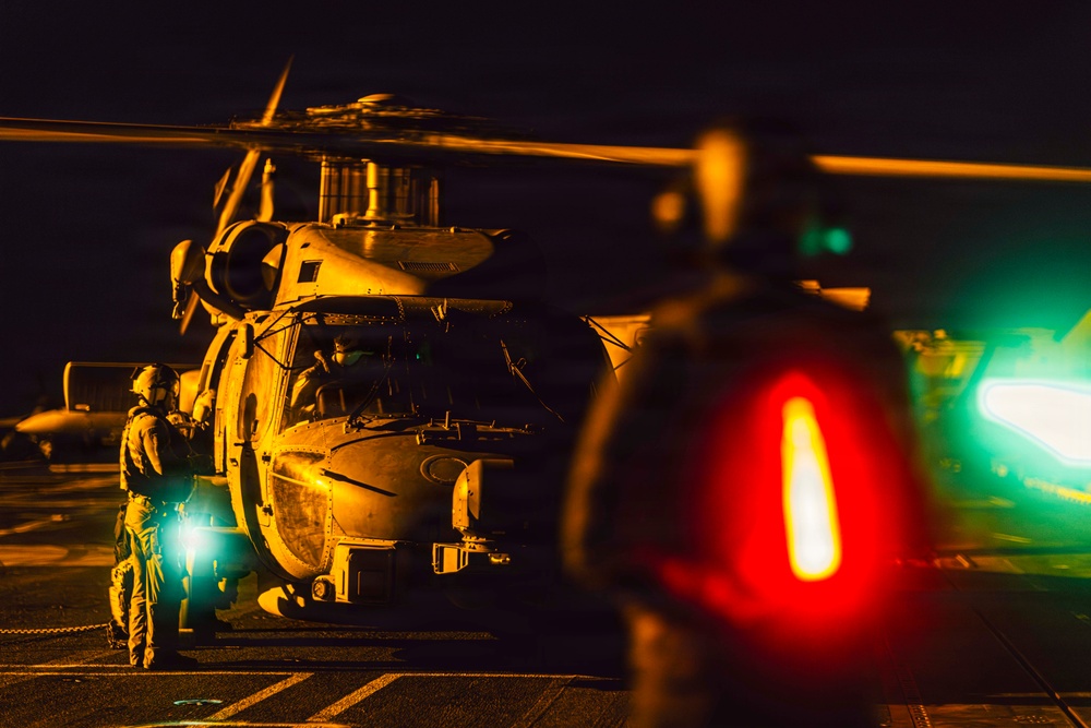 A nighttime military helicopter operations scene, likely on a naval vessel flight deck. A large military helicopter, consistent with an MH-60 Seahawk type, is on the deck with rotor blades in motion, attended by at least one ground crew member in flight gear. The scene is illuminated by a combination of warm orange ambient light, a bright white work light, and red and green navigation/signal lights in the foreground.