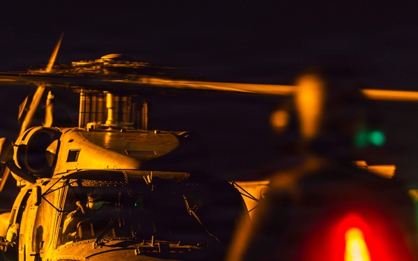 A nighttime military helicopter operations scene, likely on a naval vessel flight deck. A large military helicopter, consistent with an MH-60 Seahawk type, is on the deck with rotor blades in motion, attended by at least one ground crew member in flight gear. The scene is illuminated by a combination of warm orange ambient light, a bright white work light, and red and green navigation/signal lights in the foreground.