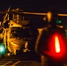 A nighttime military helicopter operations scene, likely on a naval vessel flight deck. A large military helicopter, consistent with an MH-60 Seahawk type, is on the deck with rotor blades in motion, attended by at least one ground crew member in flight gear. The scene is illuminated by a combination of warm orange ambient light, a bright white work light, and red and green navigation/signal lights in the foreground.