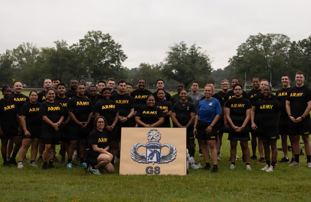 A large group of U.S. Army personnel pose for a formal group photograph on a grass field. The majority are wearing matching black 'ARMY' PT (Physical Training) shirts, with one individual in a blue shirt. They are gathered around a sign displaying the 18th Airborne Corps insignia with 'G8' designation.