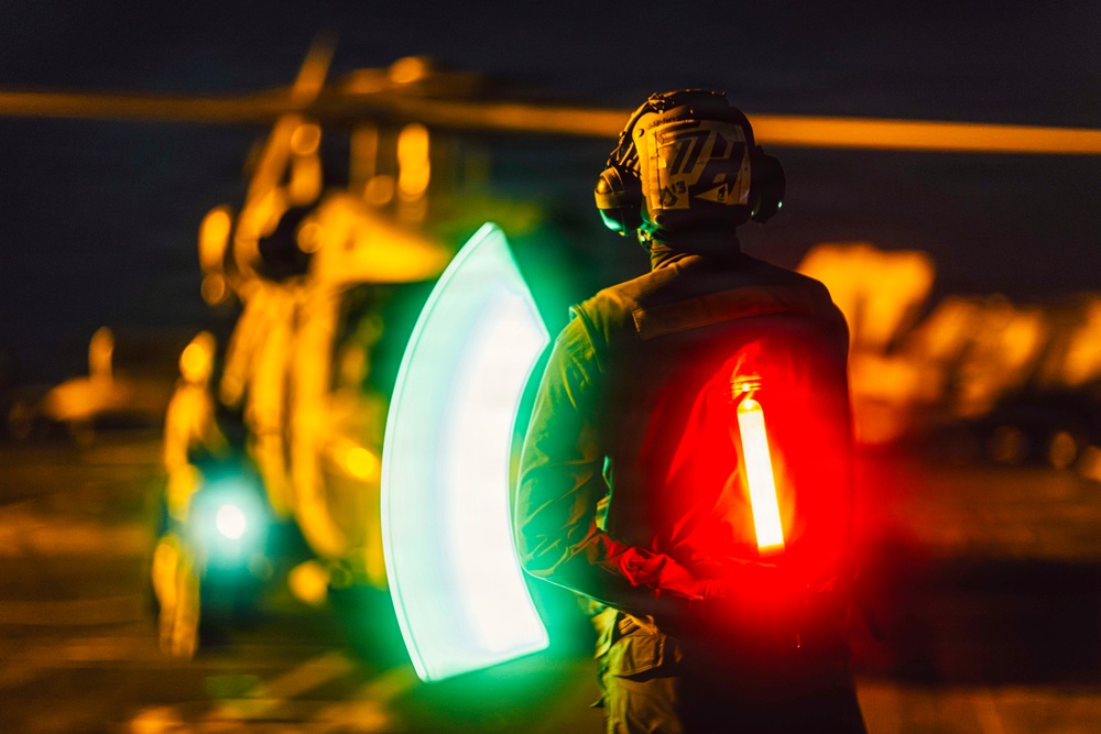 A military flight deck crew member is captured at night using illuminated wands to guide a helicopter during flight operations. The person is wearing a helmet with communications headset and holding two light wands — one green and one red — creating a motion-blur arc effect from the sweeping movement. A military helicopter is visible in the blurred background, illuminated by warm orange lighting.