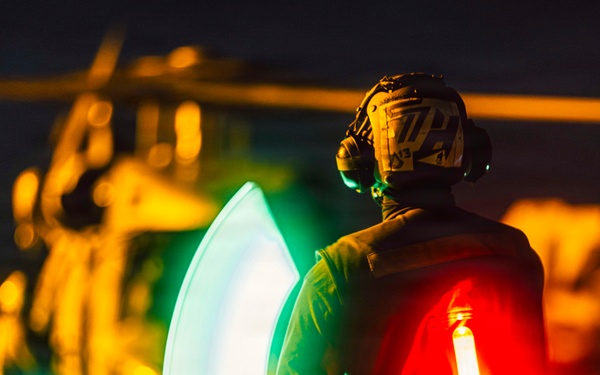 A military flight deck crew member is captured at night using illuminated wands to guide a helicopter during flight operations. The person is wearing a helmet with communications headset and holding two light wands — one green and one red — creating a motion-blur arc effect from the sweeping movement. A military helicopter is visible in the blurred background, illuminated by warm orange lighting.
