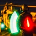 A military flight deck crew member is captured at night using illuminated wands to guide a helicopter during flight operations. The person is wearing a helmet with communications headset and holding two light wands — one green and one red — creating a motion-blur arc effect from the sweeping movement. A military helicopter is visible in the blurred background, illuminated by warm orange lighting.