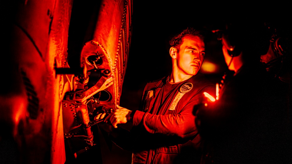A technician or military maintenance personnel is working on mechanical components of what appears to be an aircraft or helicopter under red-tinted lighting at night. The individual is using a tool, likely a wrench, to perform maintenance on riveted metal panels and mechanical assemblies. A second partially visible person is present to the right, also engaged in the work.