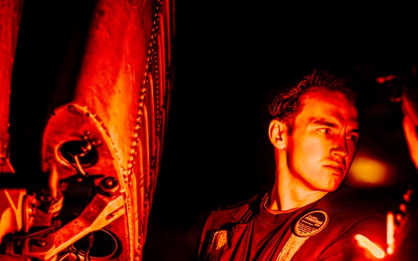 A technician or military maintenance personnel is working on mechanical components of what appears to be an aircraft or helicopter under red-tinted lighting at night. The individual is using a tool, likely a wrench, to perform maintenance on riveted metal panels and mechanical assemblies. A second partially visible person is present to the right, also engaged in the work.