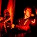 A technician or military maintenance personnel is working on mechanical components of what appears to be an aircraft or helicopter under red-tinted lighting at night. The individual is using a tool, likely a wrench, to perform maintenance on riveted metal panels and mechanical assemblies. A second partially visible person is present to the right, also engaged in the work.