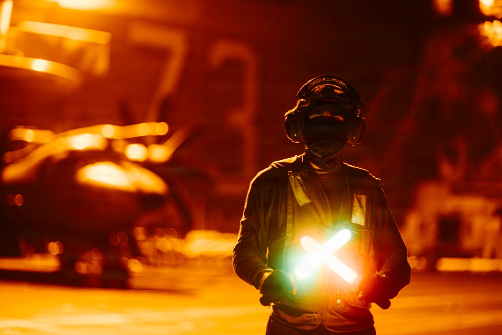 A military flight deck crew member stands on what appears to be an aircraft carrier flight deck during nighttime operations, illuminated by intense orange ambient lighting. The individual is holding two crossed luminescent wands forming an 'X' shape, a standard signal used in aircraft marshalling. The background shows blurred aircraft and deck equipment bathed in warm orange light.