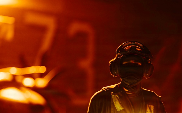 A military flight deck crew member stands on what appears to be an aircraft carrier flight deck during nighttime operations, illuminated by intense orange ambient lighting. The individual is holding two crossed luminescent wands forming an 'X' shape, a standard signal used in aircraft marshalling. The background shows blurred aircraft and deck equipment bathed in warm orange light.