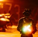 A military flight deck crew member stands on what appears to be an aircraft carrier flight deck during nighttime operations, illuminated by intense orange ambient lighting. The individual is holding two crossed luminescent wands forming an 'X' shape, a standard signal used in aircraft marshalling. The background shows blurred aircraft and deck equipment bathed in warm orange light.