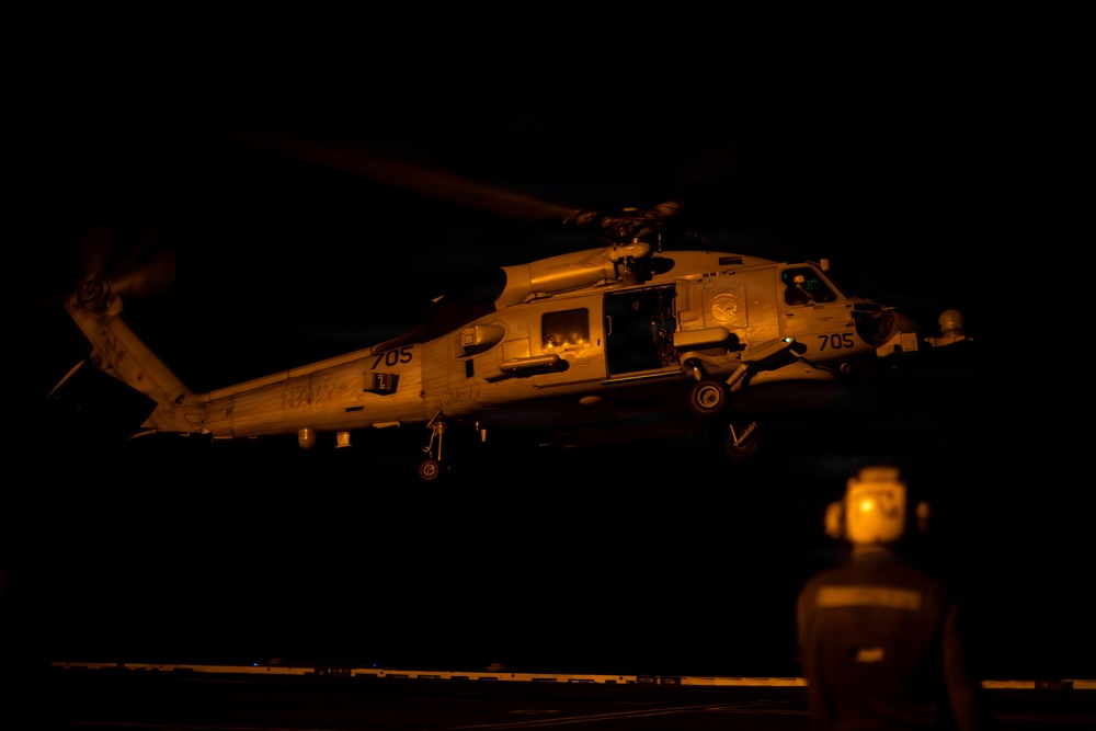 A U.S. Navy helicopter, marked with the number '705' and the word 'NAVY,' is captured during a nighttime operation, likely hovering or landing on a ship's flight deck. The scene is illuminated by warm orange ambient lighting, revealing ground crew personnel observing the aircraft. The helicopter's cabin door is open, suggesting active crew operations or personnel transfer.