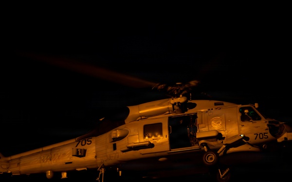 A U.S. Navy helicopter, marked with the number '705' and the word 'NAVY,' is captured during a nighttime operation, likely hovering or landing on a ship's flight deck. The scene is illuminated by warm orange ambient lighting, revealing ground crew personnel observing the aircraft. The helicopter's cabin door is open, suggesting active crew operations or personnel transfer.
