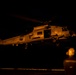 A U.S. Navy helicopter, marked with the number '705' and the word 'NAVY,' is captured during a nighttime operation, likely hovering or landing on a ship's flight deck. The scene is illuminated by warm orange ambient lighting, revealing ground crew personnel observing the aircraft. The helicopter's cabin door is open, suggesting active crew operations or personnel transfer.