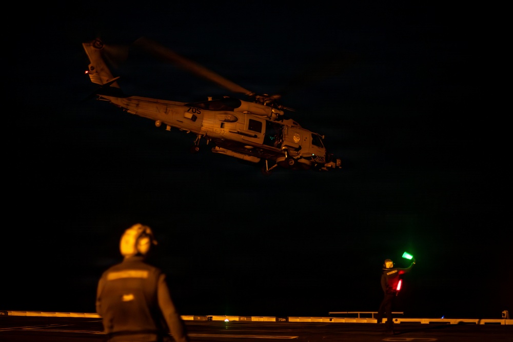 A military helicopter, marked with the number '705', is captured during a nighttime flight deck operation, likely aboard a naval vessel. Two deck crew members are visible on the flight deck, one using illuminated signal wands (red and green lights) to guide the aircraft. The scene is predominantly dark with the helicopter and personnel illuminated by ambient and signal lighting.