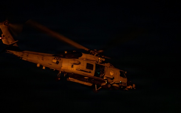 A military helicopter, marked with the number '705', is captured during a nighttime flight deck operation, likely aboard a naval vessel. Two deck crew members are visible on the flight deck, one using illuminated signal wands (red and green lights) to guide the aircraft. The scene is predominantly dark with the helicopter and personnel illuminated by ambient and signal lighting.