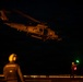 A military helicopter, marked with the number '705', is captured during a nighttime flight deck operation, likely aboard a naval vessel. Two deck crew members are visible on the flight deck, one using illuminated signal wands (red and green lights) to guide the aircraft. The scene is predominantly dark with the helicopter and personnel illuminated by ambient and signal lighting.