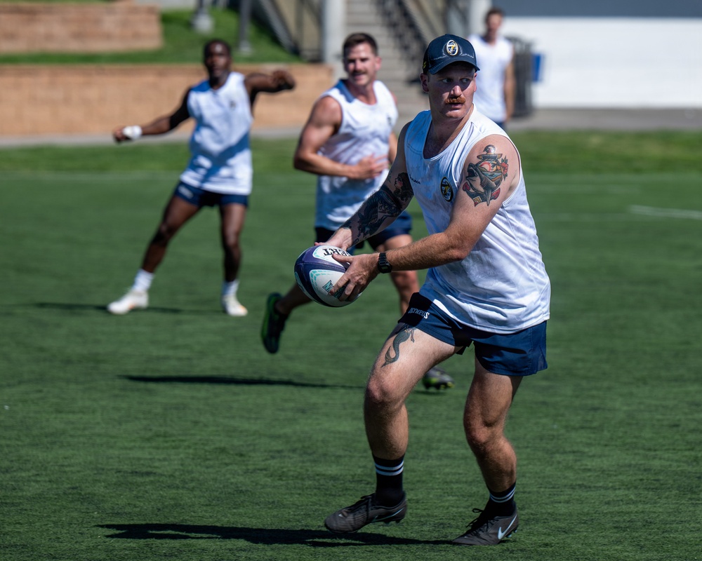 A group of rugby players are engaged in a training session on a well-maintained grass field. The foreground player, heavily tattooed and wearing a navy cap, is in the act of passing or carrying a rugby ball. Several teammates in matching white and navy uniforms are visible in the background.