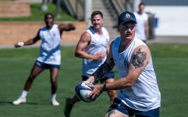 A group of rugby players are engaged in a training session on a well-maintained grass field. The foreground player, heavily tattooed and wearing a navy cap, is in the act of passing or carrying a rugby ball. Several teammates in matching white and navy uniforms are visible in the background.