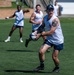 A group of rugby players are engaged in a training session on a well-maintained grass field. The foreground player, heavily tattooed and wearing a navy cap, is in the act of passing or carrying a rugby ball. Several teammates in matching white and navy uniforms are visible in the background.