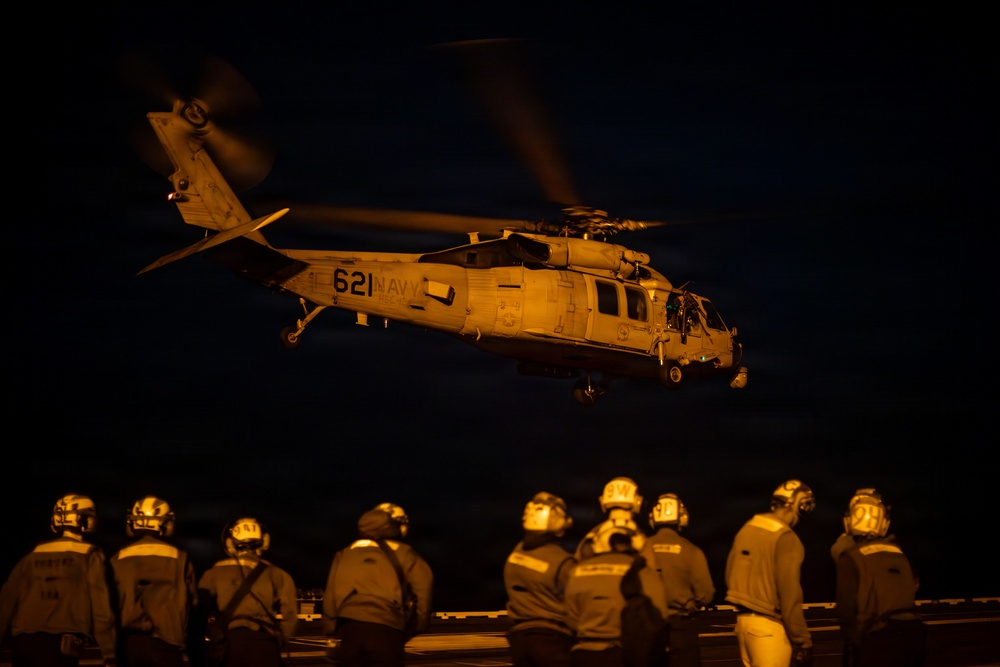 A U.S. Navy helicopter marked '621 NAVY' is captured during a nighttime flight operation above what appears to be the deck of an aircraft carrier or naval vessel. Multiple flight deck crew members wearing helmets and protective gear observe the helicopter from below, illuminated by warm amber deck lighting. The scene depicts a nighttime carrier aviation operation with ground crew standing by.