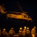 A U.S. Navy helicopter marked '621 NAVY' is captured during a nighttime flight operation above what appears to be the deck of an aircraft carrier or naval vessel. Multiple flight deck crew members wearing helmets and protective gear observe the helicopter from below, illuminated by warm amber deck lighting. The scene depicts a nighttime carrier aviation operation with ground crew standing by.