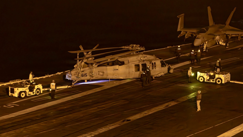 A nighttime scene on a U.S. Navy aircraft carrier flight deck showing a military helicopter numbered '615' being positioned or serviced by deck crew. Multiple ground support vehicles and personnel are visible around the helicopter, with a fixed-wing jet aircraft (likely an F/A-18) visible in the background. The scene is illuminated by artificial deck lighting, casting a warm amber glow across the operations.