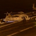 A nighttime scene on a U.S. Navy aircraft carrier flight deck showing a military helicopter numbered '615' being positioned or serviced by deck crew. Multiple ground support vehicles and personnel are visible around the helicopter, with a fixed-wing jet aircraft (likely an F/A-18) visible in the background. The scene is illuminated by artificial deck lighting, casting a warm amber glow across the operations.
