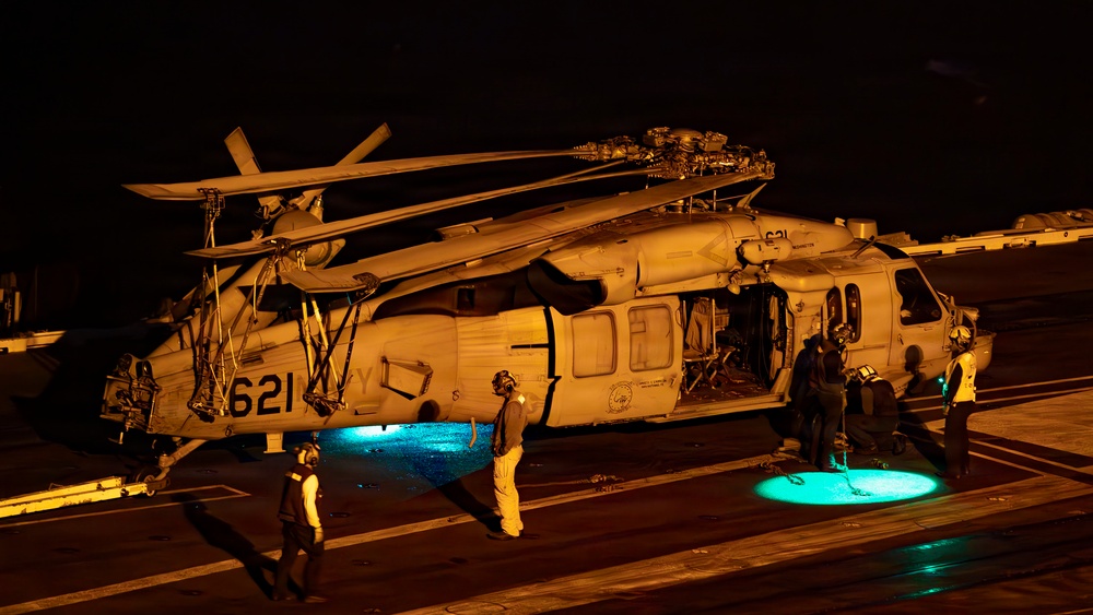 A U.S. Navy helicopter, marked with the number '621' and partially visible 'NAVY' text, is parked on what appears to be an aircraft carrier flight deck at night. The scene is illuminated by warm orange artificial lighting and two teal/cyan ground lights beneath the aircraft. Several crew members in flight deck gear are gathered around the helicopter, apparently conducting maintenance or pre/post-flight operations.