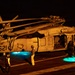 A U.S. Navy helicopter, marked with the number '621' and partially visible 'NAVY' text, is parked on what appears to be an aircraft carrier flight deck at night. The scene is illuminated by warm orange artificial lighting and two teal/cyan ground lights beneath the aircraft. Several crew members in flight deck gear are gathered around the helicopter, apparently conducting maintenance or pre/post-flight operations.