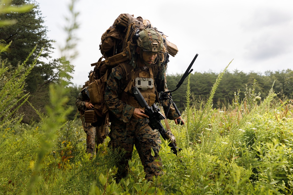A U.S. Marine in full combat gear moves through dense vegetation, carrying a heavily loaded rucksack with camouflage netting and holding an assault rifle. A second Marine is visible in the background. The setting appears to be a field training exercise in a forested, overgrown area.