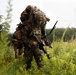 A U.S. Marine in full combat gear moves through dense vegetation, carrying a heavily loaded rucksack with camouflage netting and holding an assault rifle. A second Marine is visible in the background. The setting appears to be a field training exercise in a forested, overgrown area.