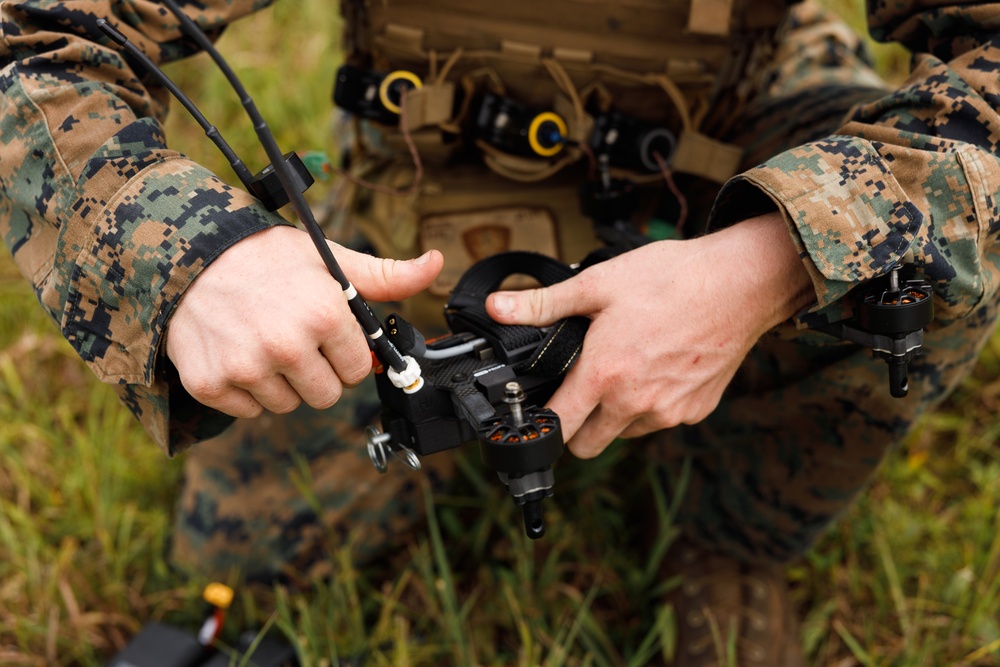 A military personnel wearing MARPAT (Marine pattern) digital camouflage uniform is handling a small FPV or racing-style drone with carbon fiber frame components. The individual appears to be assembling, inspecting, or preparing the drone for deployment in a field environment. The setting appears to be an outdoor grassy area during what looks like a training or operational exercise.