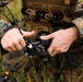 A military personnel wearing MARPAT (Marine pattern) digital camouflage uniform is handling a small FPV or racing-style drone with carbon fiber frame components. The individual appears to be assembling, inspecting, or preparing the drone for deployment in a field environment. The setting appears to be an outdoor grassy area during what looks like a training or operational exercise.