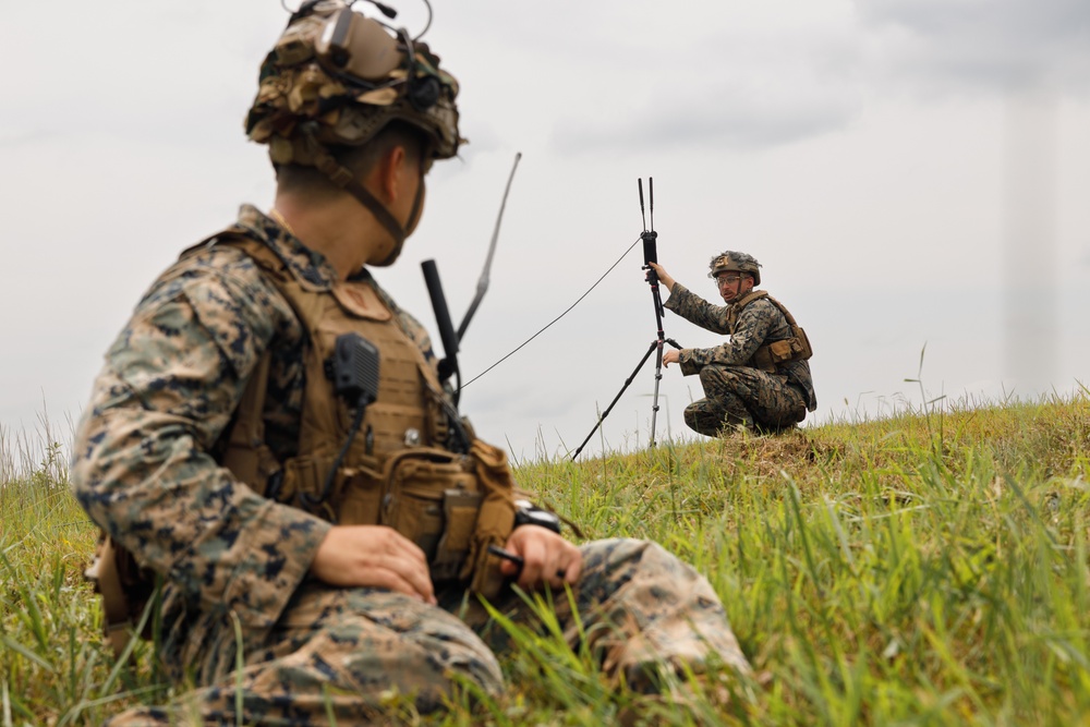 Two military personnel in MARPAT (Marine pattern) camouflage uniforms are positioned in an open grassy field under overcast skies. The foreground soldier is seated and equipped with tactical gear and a radio, while the background soldier is crouching and setting up what appears to be a tripod-mounted antenna or communications device. The scene suggests a field communications exercise or operational communications setup.