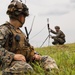 Two military personnel in MARPAT (Marine pattern) camouflage uniforms are positioned in an open grassy field under overcast skies. The foreground soldier is seated and equipped with tactical gear and a radio, while the background soldier is crouching and setting up what appears to be a tripod-mounted antenna or communications device. The scene suggests a field communications exercise or operational communications setup.
