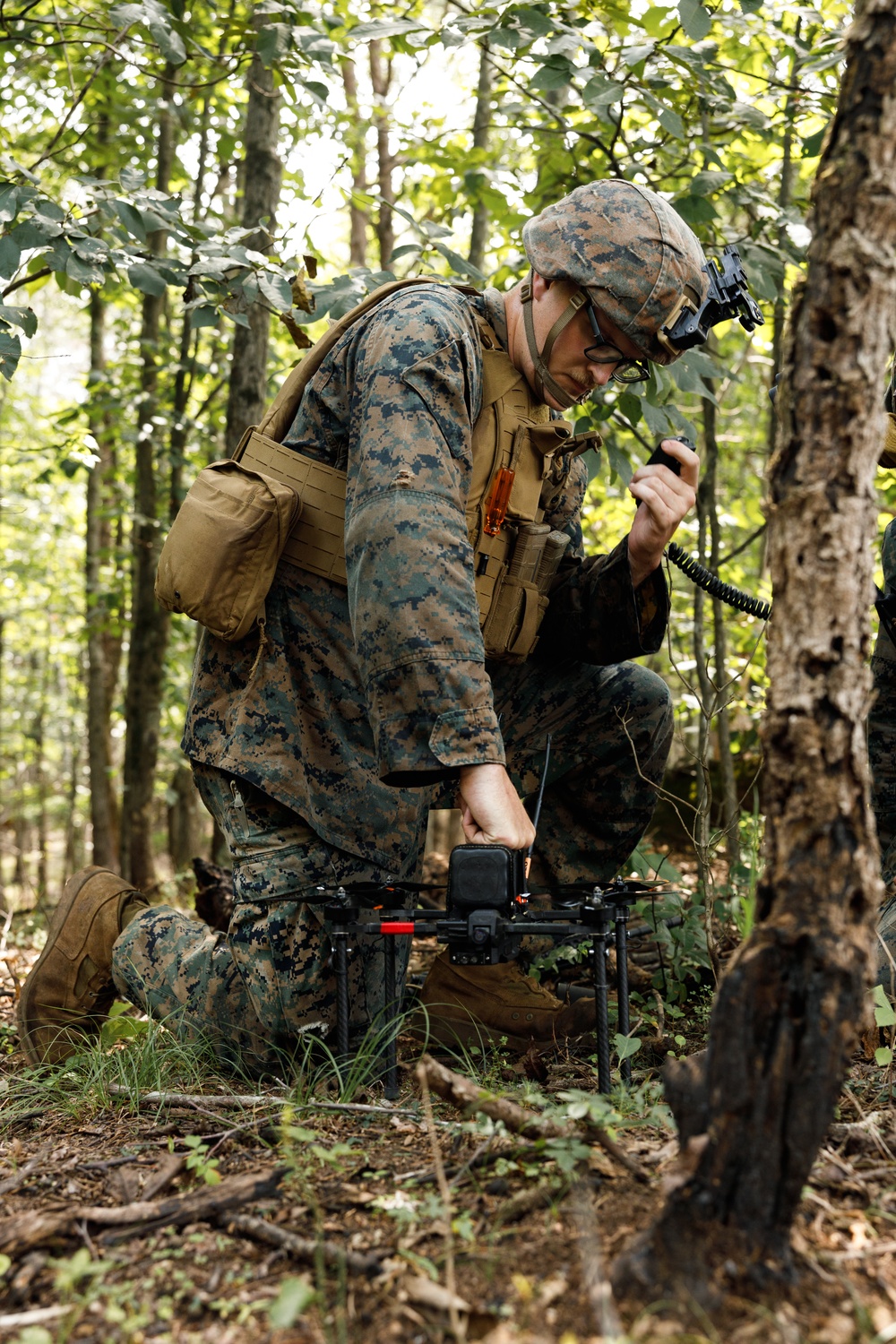 A military service member in MARPAT (Marine pattern) camouflage uniform is kneeling in a wooded area while handling a small multi-rotor drone on the ground. The individual is simultaneously holding what appears to be a radio handset, suggesting communication during drone preparation or operation. The setting is a deciduous forest with dense foliage, indicating a field training or operational environment.
