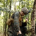 A military service member in MARPAT (Marine pattern) camouflage uniform is kneeling in a wooded area while handling a small multi-rotor drone on the ground. The individual is simultaneously holding what appears to be a radio handset, suggesting communication during drone preparation or operation. The setting is a deciduous forest with dense foliage, indicating a field training or operational environment.