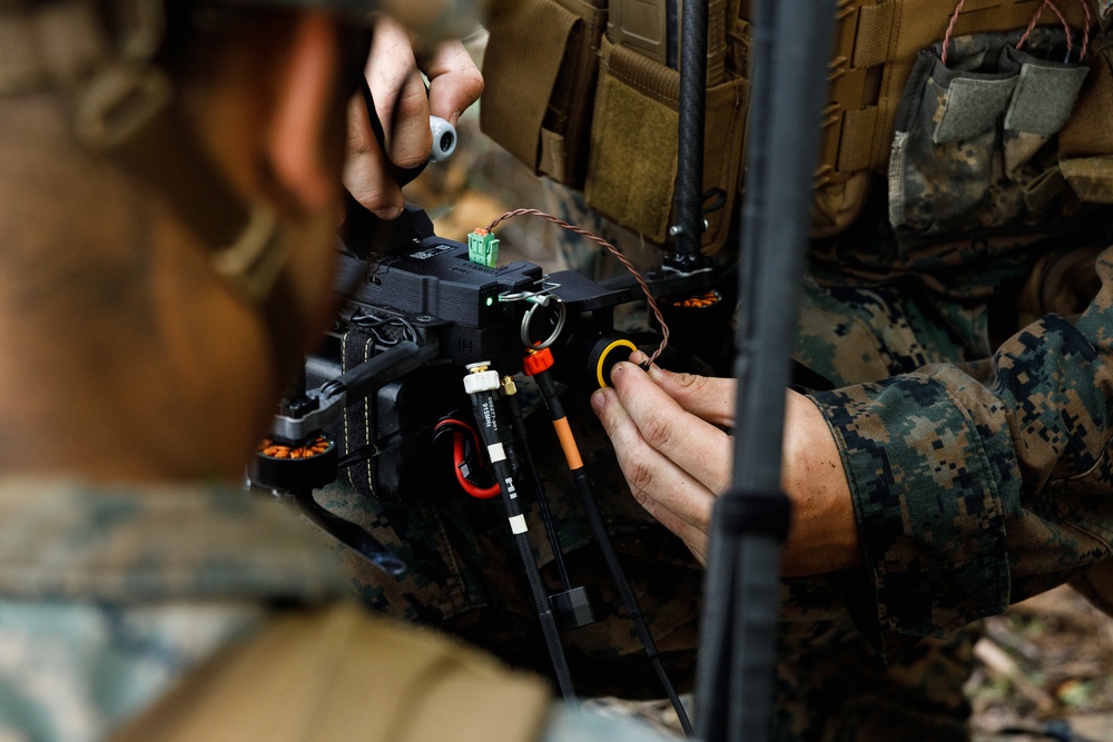 Two military personnel in MARPAT (Marine digital camouflage) uniforms are working on what appears to be a small unmanned aerial vehicle (drone) or electronic equipment assembly. They are connecting wiring and components, with visible colored cables (red, black, orange) and electronic connectors. The setting appears to be an outdoor wooded environment, suggesting field operations or training.