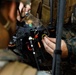 Two military personnel in MARPAT (Marine digital camouflage) uniforms are working on what appears to be a small unmanned aerial vehicle (drone) or electronic equipment assembly. They are connecting wiring and components, with visible colored cables (red, black, orange) and electronic connectors. The setting appears to be an outdoor wooded environment, suggesting field operations or training.