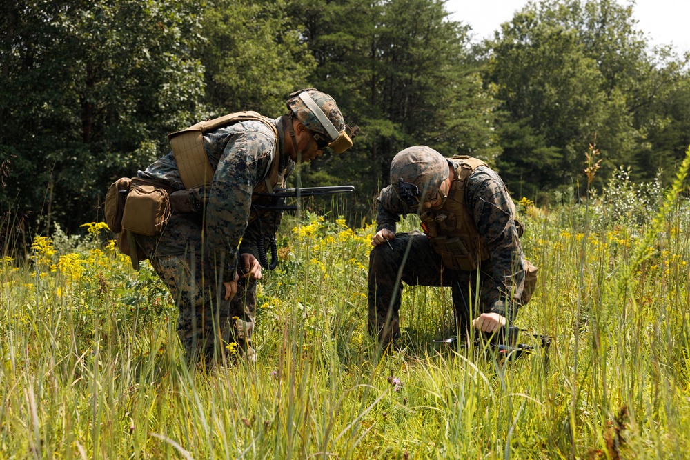 Two military personnel in U.S. Marine Corps MARPAT camouflage uniforms are crouched in an open field examining an object on the ground. They are equipped with tactical gear including helmets, plate carriers, and backpacks. The setting appears to be a training exercise in a wooded, grassy environment during summer.