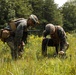 Two military personnel in U.S. Marine Corps MARPAT camouflage uniforms are crouched in an open field examining an object on the ground. They are equipped with tactical gear including helmets, plate carriers, and backpacks. The setting appears to be a training exercise in a wooded, grassy environment during summer.