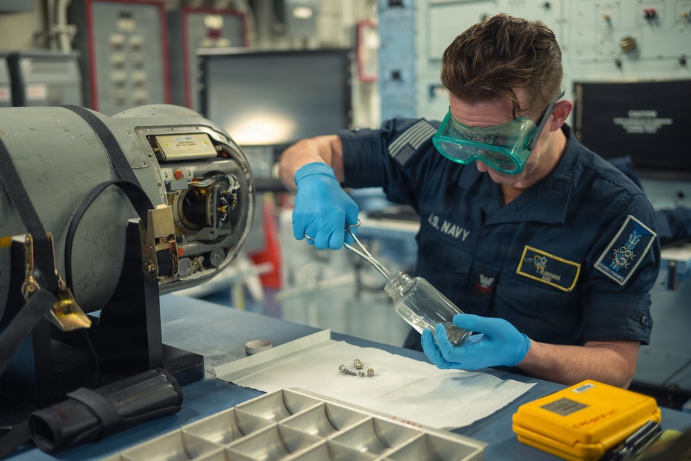 A U.S. Navy sailor wearing blue nitrile gloves and green safety goggles is performing a technical maintenance or cleaning procedure at a workbench. The sailor is using tweezers or tongs to handle small metal components, possibly placing or removing them from a glass container. The setting appears to be an onboard workshop or maintenance bay on a naval vessel.