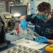 A U.S. Navy sailor wearing blue nitrile gloves and green safety goggles is performing a technical maintenance or cleaning procedure at a workbench. The sailor is using tweezers or tongs to handle small metal components, possibly placing or removing them from a glass container. The setting appears to be an onboard workshop or maintenance bay on a naval vessel.