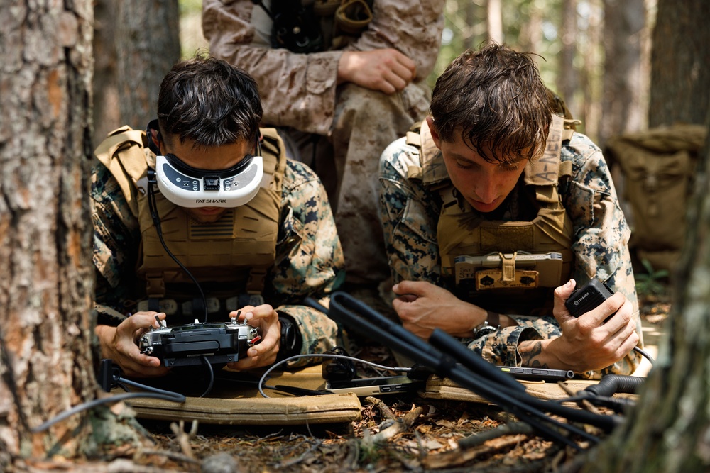 Two military personnel in MARPAT digital camouflage uniforms are operating FPV (First Person View) drone equipment in a wooded outdoor setting. The left individual is wearing Fat Shark FPV goggles and holding a drone controller, while the right individual is monitoring a tablet or display device. A third uniformed individual is partially visible in the background.