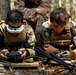 Two military personnel in MARPAT digital camouflage uniforms are operating FPV (First Person View) drone equipment in a wooded outdoor setting. The left individual is wearing Fat Shark FPV goggles and holding a drone controller, while the right individual is monitoring a tablet or display device. A third uniformed individual is partially visible in the background.