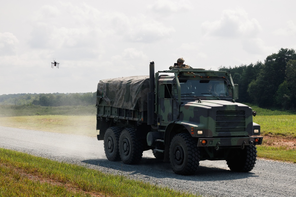 A camouflage-painted military cargo truck, consistent with a Medium Tactical Vehicle Replacement (MTVR) or similar 6x6 heavy truck, is traveling along a gravel road in what appears to be a training or exercise environment. A soldier is positioned in the turret hatch on top of the cab. A small quadcopter drone is visible in the air to the left of the vehicle.