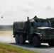 A camouflage-painted military cargo truck, consistent with a Medium Tactical Vehicle Replacement (MTVR) or similar 6x6 heavy truck, is traveling along a gravel road in what appears to be a training or exercise environment. A soldier is positioned in the turret hatch on top of the cab. A small quadcopter drone is visible in the air to the left of the vehicle.